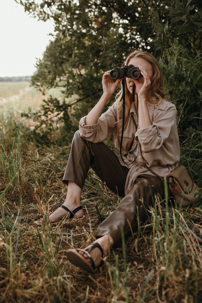 a beautiful young woman on safari sits in the grass and looks through binoculars